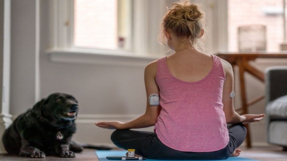A seated woman wearing medical devices on her arms performing yoga next to a dog.